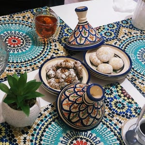 May include: A table setting featuring a patterned tablecloth with a teal, blue, and red design. It includes small ceramic bowls with cookies, a glass of tea, and decorative ceramic tagine pots with blue and white patterns. A small potted succulent is also present.