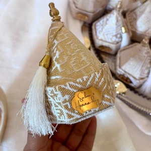 May include: A gold and white fabric cone-shaped favor box with a tassel and a gold label that reads "Salma's Henna day". The box has a textured pattern and is part of a set of similar boxes.