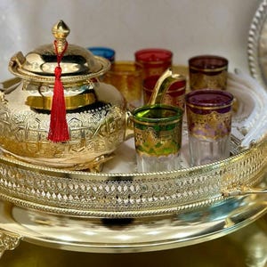 May include: A gold-colored tea set with intricate patterns, including a teapot with a red tassel and several colorful glasses. The set is displayed on a decorative gold tray, creating an elegant presentation.