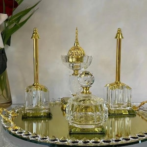 May include: A decorative set of crystal and gold-toned accessories, including a tray, two candle holders, a small jar, and a decorative piece. The items are arranged on a mirrored tray, with a vase of white roses in the background.