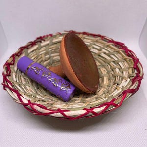 May include: A small, woven basket with a red trim holds a purple tube with gold lettering and a brown, wooden scoop. The basket is made of natural fibers and is set against a white background.