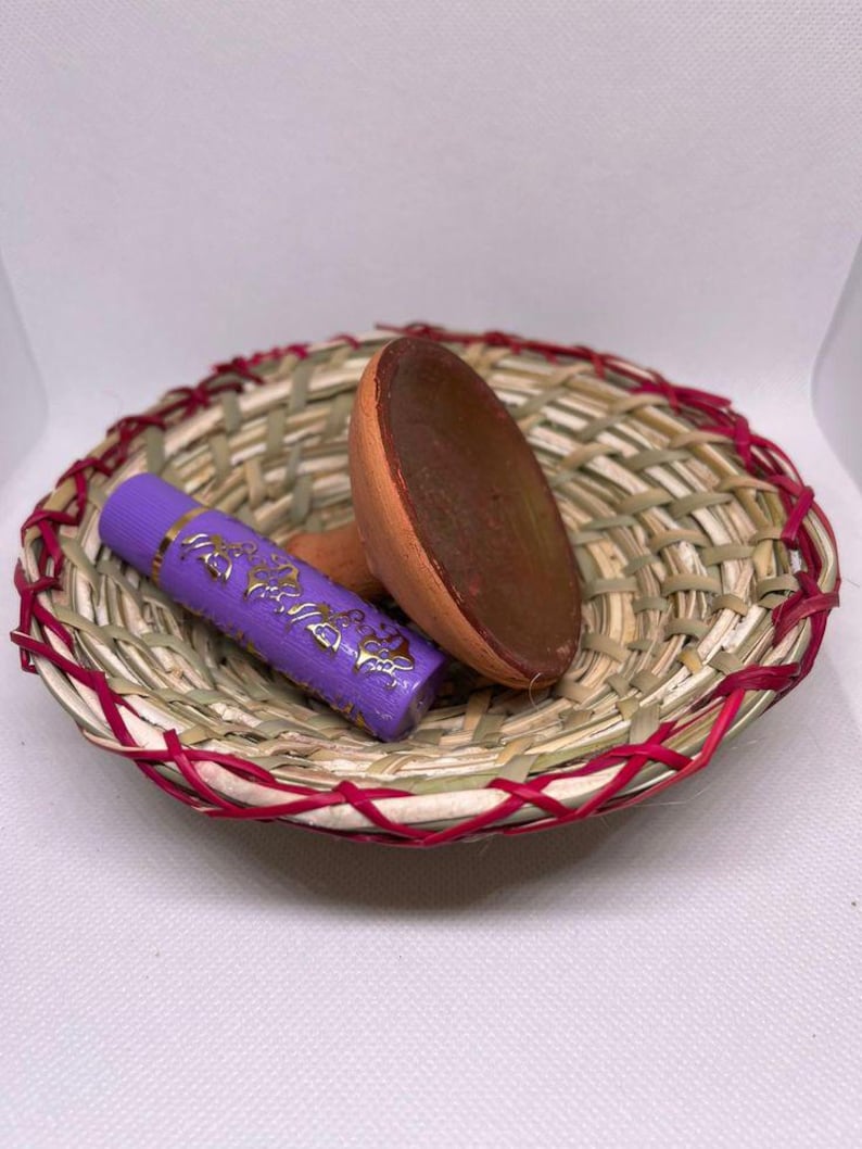 May include: A small, woven basket with a red trim holds a purple tube with gold lettering and a brown, wooden scoop. The basket is made of natural fibers and is set against a white background.