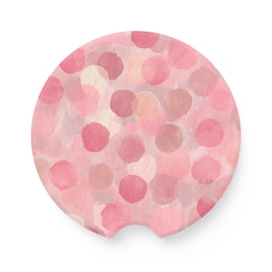 May include: A round car coaster with a watercolor design. The coaster features a pattern of pink and peach-colored polka dots on a light pink background. The coaster has a small notch on one side for easy removal from the car's cup holder.