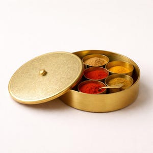 Op de afbeelding: Een goudkleurige kruidenbox met een decoratief deksel, open om zes kleine, ronde bakjes gevuld met diverse kruiden te onthullen. Een klein lepeltje rust in een van de bakjes. De box staat op een witte achtergrond.