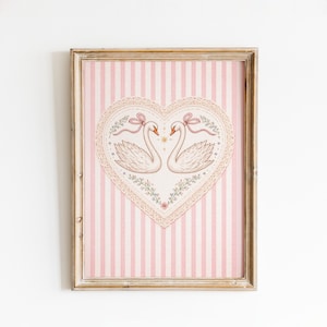 May include: Framed artwork featuring two swans facing each other within a heart-shaped design. The swans have pink bows and are surrounded by floral accents. The background has pink and white vertical stripes.