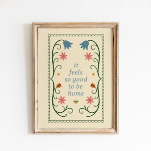 Könnte beinhalten: Gerahmter Kunstdruck mit cremefarbenem Hintergrund und dekorativem Rand. Der Druck zeigt den Text "it feels so good to be home", umgeben von floralen Illustrationen in Blau, Rosa und Orange. Der Rahmen ist hellbraun.