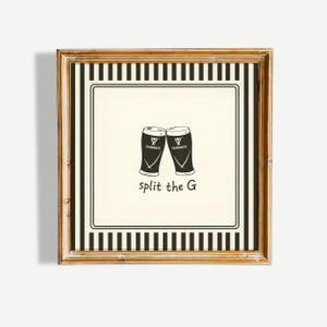 May include: A framed print with a gold-coloured frame. The print features two Guinness pint glasses clinking, with the words "split the G" below. The background is cream, and the frame has a black and white striped border.