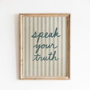 May include: A framed print with a light wood frame. The artwork features vertical stripes in cream and light blue. The words "speak your truth" are written in a teal script font.