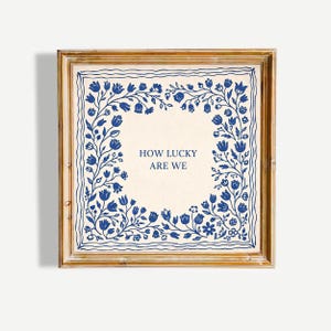 May include: A framed print with the text "HOW LUCKY ARE WE" in a square floral design. The artwork features blue flowers and leaves on a cream background, framed in a gold-toned wooden frame.