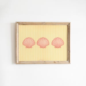 May include: A framed print with three pink scallop shells on a yellow striped background. The shells are evenly spaced and the frame is light brown. The artwork is displayed against a white wall.