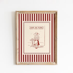 May include: Framed print featuring a vintage illustration of a person reading a newspaper at a cafe table. The print has a cream background with a red and white striped border and the words "Cafe de Flore" at the top.