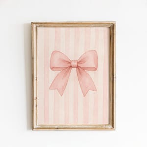 May include: A framed watercolour print showing a large, soft pink bow against a backdrop of vertical pink and white stripes. The artwork is in a gold-toned wooden frame, creating a delicate and feminine aesthetic.