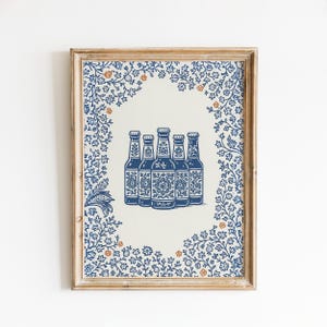 May include: Framed print featuring five blue and white beer bottles with floral designs. The bottles are centered on a cream background, surrounded by a blue floral border with orange accents. The frame is a light wood color.