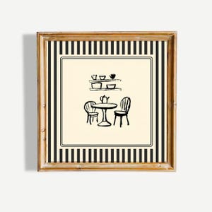 May include: A framed artwork featuring a black and white illustration of a cafe scene. The image depicts a small table with two chairs, a teapot, and a shelf with dishes. The frame has a gold finish with black and white striped borders.