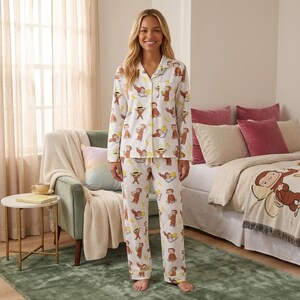 May include: A white long-sleeve button-down pajama shirt and matching pajama pants featuring an all-over print of Curious George in various poses, including wearing a yellow hat, holding a banana, and reading a book, with yellow piping trim.