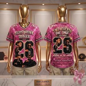 May include: A pink baseball jersey with the text "CHRIS BROWN USHER TOUR" and the number "26" on the front. The back of the jersey reads "TWO LEGENDS ONE STAGE" and "HISTORY IN THE MAKING". The jersey is displayed on a mannequin.