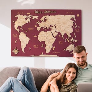 May include: A burgundy wooden world map with the text "Oliver's Adventures" hangs on a wall. The map features detailed country outlines in a lighter wood tone. A couple is seated on a gray sofa, looking at a laptop.