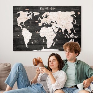 Peut inclure: Une carte du monde en bois noir avec le texte "Our Adventure" est accrochée au mur au-dessus d'un couple assis sur un canapé beige. La carte a un contour clair du monde. Le couple est habillé décontracté, savourant une boisson.