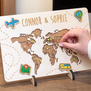 May include: A wooden world map with the names "Connor & Sophie" at the top. The map features a brown world outline on a cream background, with colorful pins and travel-themed wooden cutouts like a plane, hot air balloon, map, and bus.