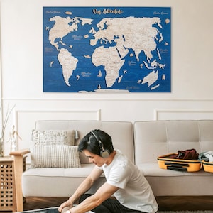 May include: A wooden world map with the text "Our Adventure" hangs on a blue wall. A person is packing a suitcase on a rug. The suitcase is white with a yellow interior. Other luggage is nearby.