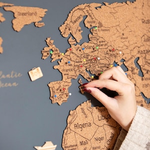 May include: A cork world map with country outlines and city names, mounted on a dark gray background. The map is decorated with colorful pins and wooden cutouts of a suitcase and boat. The words "Lucas and Emma Adventure" are above the map.