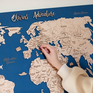 May include: A detailed wooden world map on a blue background, with the text "Olivia's Adventure". The map is being marked with a pin. Decorative elements include a plane, boat, and hot air balloon. The map is made of wood.