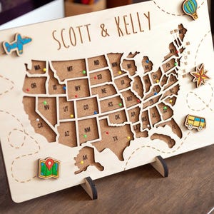 May include: Wooden map of the United States with state outlines and the names "SCOTT & KELLY" engraved at the top. Includes colorful pins and decorative wooden cutouts of a plane, hot air balloon, star, map, and bus.