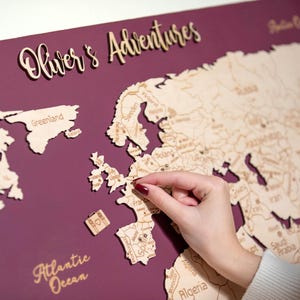 May include: A wooden world map with the text "Oliver's Adventures" in a cursive font. The map is mounted on a deep purple background. Small wooden travel-themed cutouts are included. A hand is placing a pin on the map.