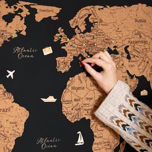 May include: A black world map with a cork overlay, featuring the text "Lucas and Emma Adventure". The map is decorated with pins and wooden cutouts of a plane, boat, suitcase, dolphin, and penguin.