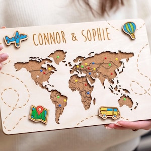 May include: A wooden world map with cut-out continents and the names "CONNOR & SOPHIE" engraved at the top. The map features colorful pins marking locations and decorative travel-themed magnets, including a plane, hot air balloon, map, and van.