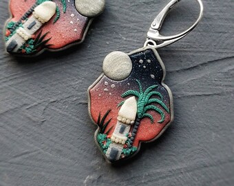 May include: A pair of dangle earrings with a silver hook and a red, black, and white clay charm featuring a palm tree, a building, and a moon.
