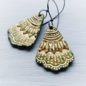 May include: A pair of green and cream ceramic earrings with a geometric design. The earrings are shaped like a teardrop and have a scalloped edge.