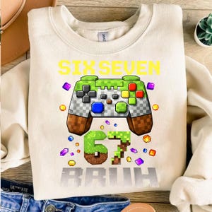 Op de afbeelding: Cr&egrave;mekleurige sweatshirt met een grote afbeelding van een gepixelde gamecontroller in groen, grijs, bruin en rood. De tekst "SIX SEVEN BRUH" staat in geel en grijs. Kleurrijke gepixelde vormen omringen de controller en de tekst.