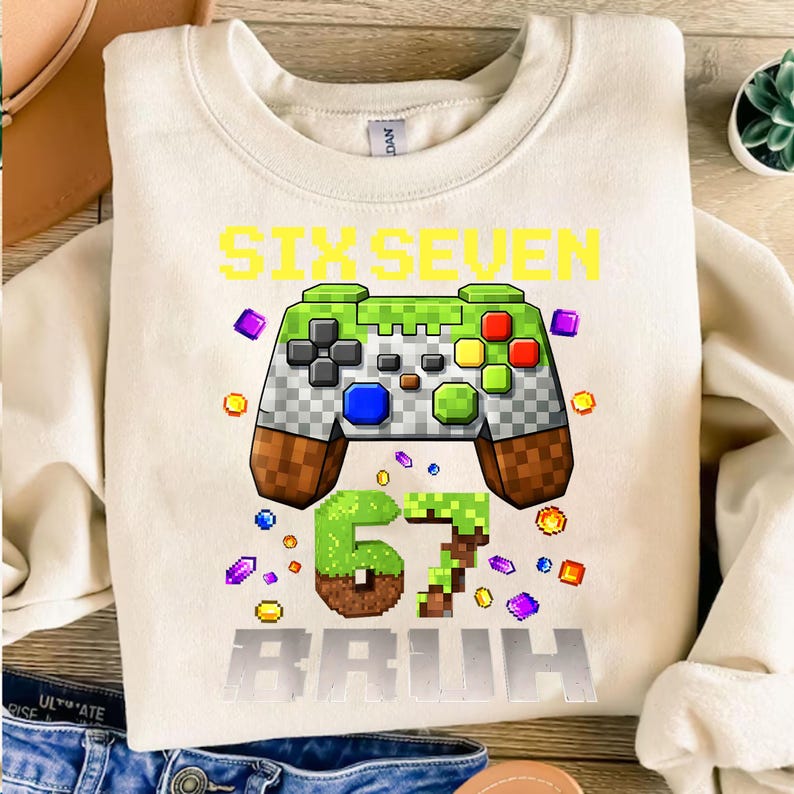 Op de afbeelding: Cr&egrave;mekleurige sweatshirt met een grote afbeelding van een gepixelde gamecontroller in groen, grijs, bruin en rood. De tekst "SIX SEVEN BRUH" staat in geel en grijs. Kleurrijke gepixelde vormen omringen de controller en de tekst.