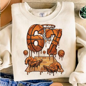 Op de afbeelding: Cr&egrave;mekleurige sweatshirt met een basketbalthema. Het ontwerp toont de nummers "67" in een druipende basketbalstijl, een basketbalveld, ballen en handen. De sweatshirt heeft een ronde hals.