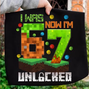 May include: Black graphic t-shirt with pixelated numbers and text. The shirt reads "I WAS 6 NOW I'M 7 UNLOCKED" in green, orange, and white. Colorful pixelated accents surround the numbers. The design is reminiscent of a popular video game.