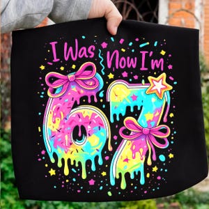 May include: Black tote bag with the text "I Was Now I'm" in pink. The number "67" is designed with a colourful, dripping, and sprinkled effect, with bows and stars. The bag is held by a person.