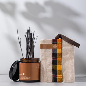 Op de afbeelding: Een bruine glazen pot met een zwart deksel, gevuld met donkere stokjes, en een beige tas met een ruitpatroon. De pot heeft een label met de tekst "ANRAT" en "MARVANILLA". De tas heeft een bruine leren riem en een ruitpatroon van geel, rood en zwart.