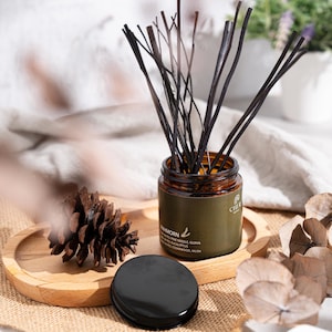 May include: A brown glass aromatherapy diffuser jar with dark reeds, a black lid, and a wooden tray. The jar has a green label with white text. A pine cone and dried leaves are also present.