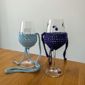 May include: Two wine glasses with crocheted cozies. One glass has a light blue cozy with a matching strap, and the other has a dark blue cozy. The glasses have decorative elements, and one has text that reads "Ahr".