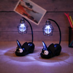 May include: A white ceramic mug with a black handle and the text "But First, Coffee" in a bold, black font. The mug is set against a blurred background, suggesting a cozy setting. The mug appears to be approximately 4 inches tall.