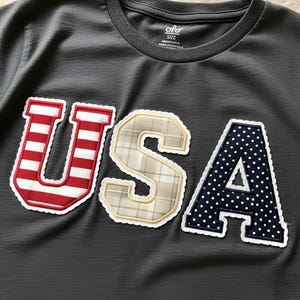 Puede incluir: Camiseta gris oscuro con la palabra "USA" aplicada en la parte delantera. La "U" es a rayas rojas y blancas, la "S" es de cuadros beige y la "A" es azul marino con lunares blancos. La camiseta tiene cuello redondo.