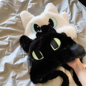 May include: Two plush cat-shaped hats, one white and one black, with large, expressive eyes. The white hat has light blue accents around the eyes, while the black hat has green accents. The hats are made of soft, fluffy material.