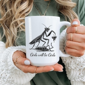 May include: A white ceramic mug featuring a black graphic illustration of two praying mantises. The design depicts one mantis standing over another with the text Girls will be Girls written in a cursive script font below the insect illustration.