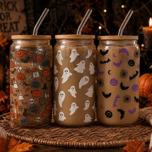 May include: Three clear glass tumblers with bamboo lids and glass straws. The first features orange and black Halloween icons, the second has white ghost silhouettes, and the third displays purple and black bats, spiders, and pumpkins.