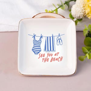 May include: A square white ceramic trinket dish with a gold-toned rim. The center features a blue graphic of a striped swimsuit, a towel, and flip-flops hanging on a clothesline, with the red text "SEE YOU AT THE BEACH" printed below.
