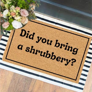May include: A rectangular tan coir doormat with a black border and the text "Did you bring a shrubbery?" in black lettering. The mat is layered over a white and black striped fabric rug, with a bouquet of pink and white flowers nearby.