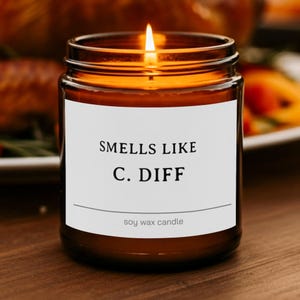 May include: A lit candle in a brown glass jar with a white label. The label features the black text SMELLS LIKE C. DIFF and the smaller text soy wax candle below a horizontal line. The flame is bright and visible at the top of the wax.
