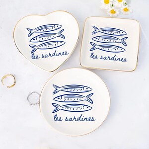 May include: A set of three white ceramic trinket dishes in heart, square, and round shapes. Each dish features a blue illustration of three sardines and the text "les sardines" in a script font. The dishes have gold rims.