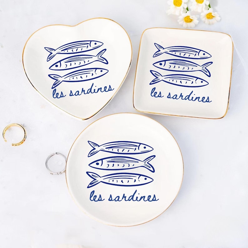May include: A set of three white ceramic trinket dishes in heart, square, and round shapes. Each dish features a blue illustration of three sardines and the text "les sardines" in a script font. The dishes have gold rims.